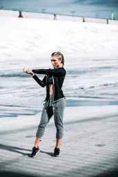 Athletic blonde woman stretching arms outdoors wearing a black jacket and grey sweatpants on a concrete spillway