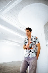 Man wearing a patterned short sleeve button shirt and grey trousers leaning against a large white structural column
