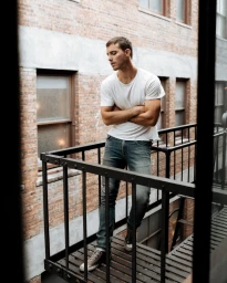 Man wearing a white t-shirt and blue jeans standing on a metal fire escape with a brick building background.
