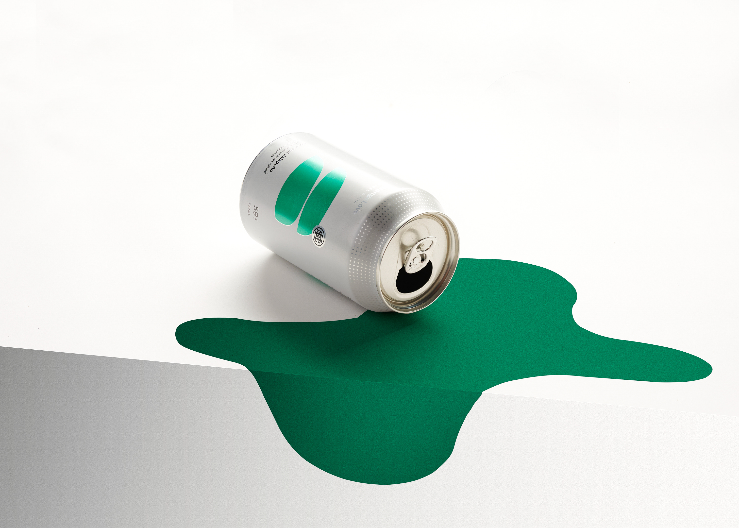 Silver beverage can lies on its side with a green liquid spill over a white table surface in a studio product shot, clean and simple.