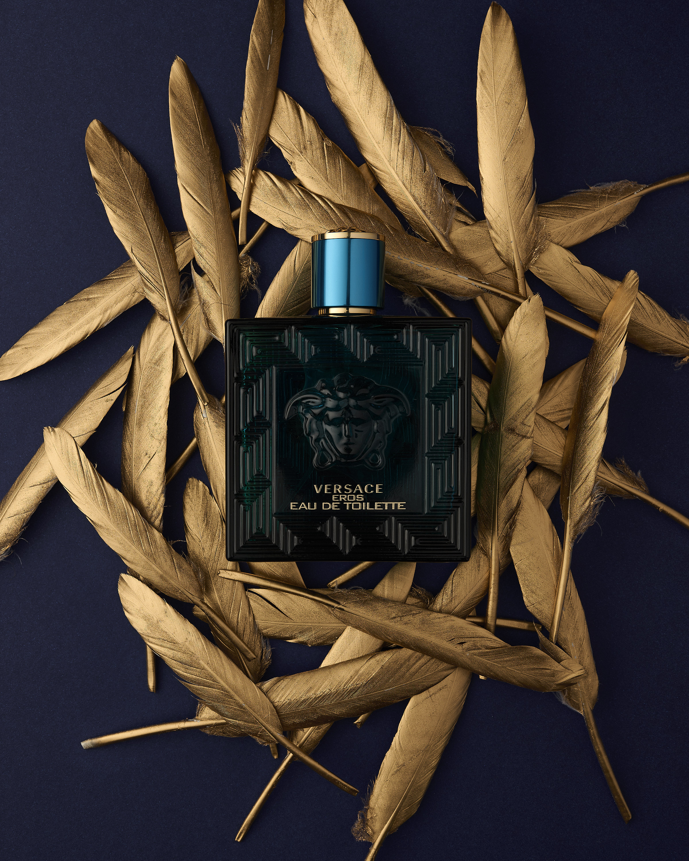 Versace Eros Eau de Toilette perfume bottle centered on navy background, surrounded by golden feathers for a luxurious, opulent feel.