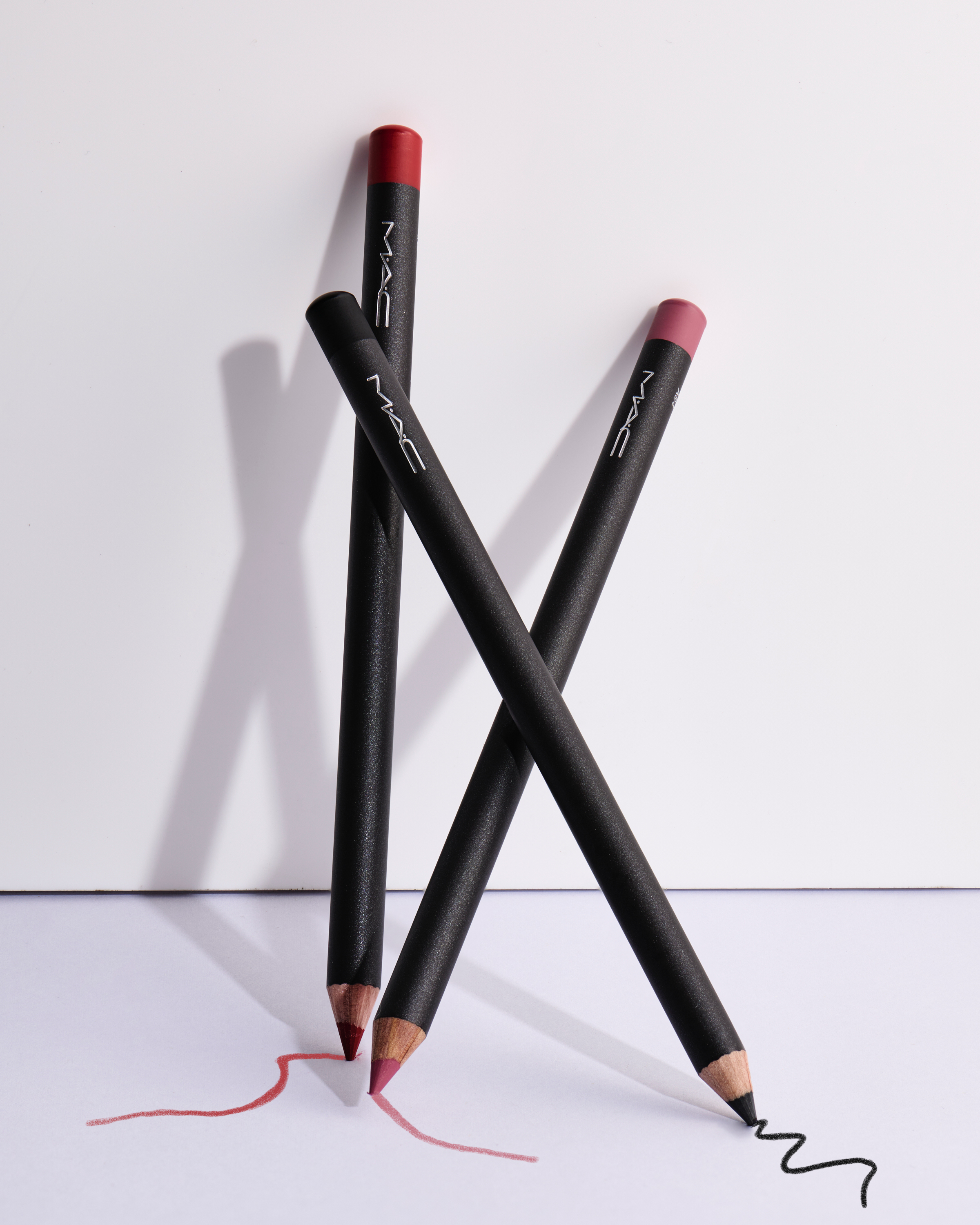Cosmetic still life featuring three MAC lip pencils with visible strokes in corresponding colors on a white surface.
