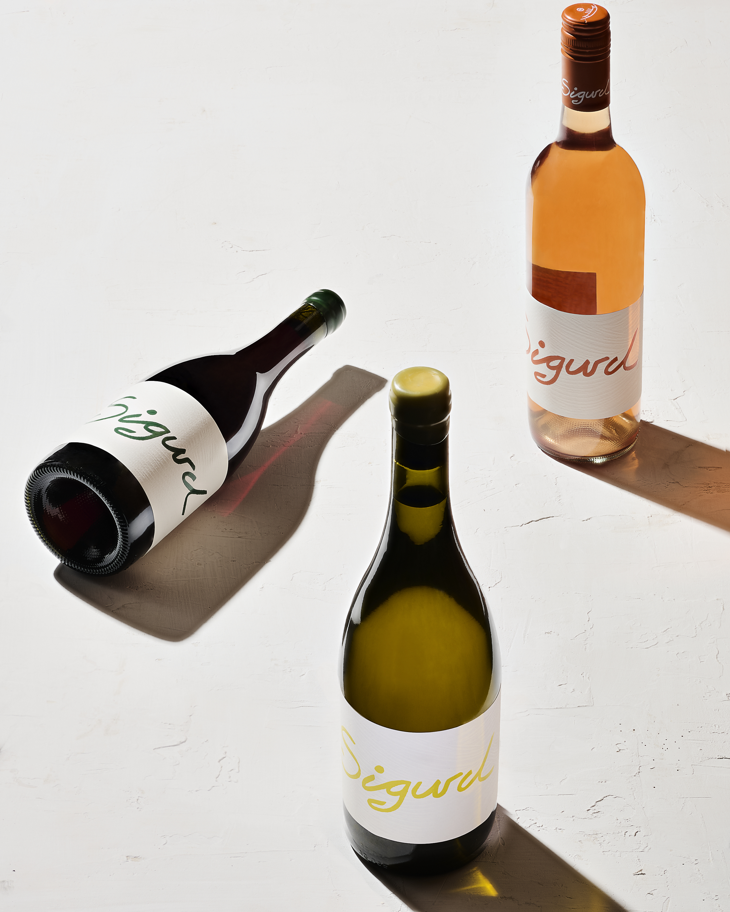 Three bottles of Sigurd wine, including a red, white, and rose, are arranged on a white background with natural lighting and shadows.