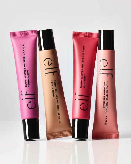 Four colorful tubes of ELF Glow Reviver Melting Lip Balm in pink, peach, and red tones against a neutral backdrop. Beauty product.