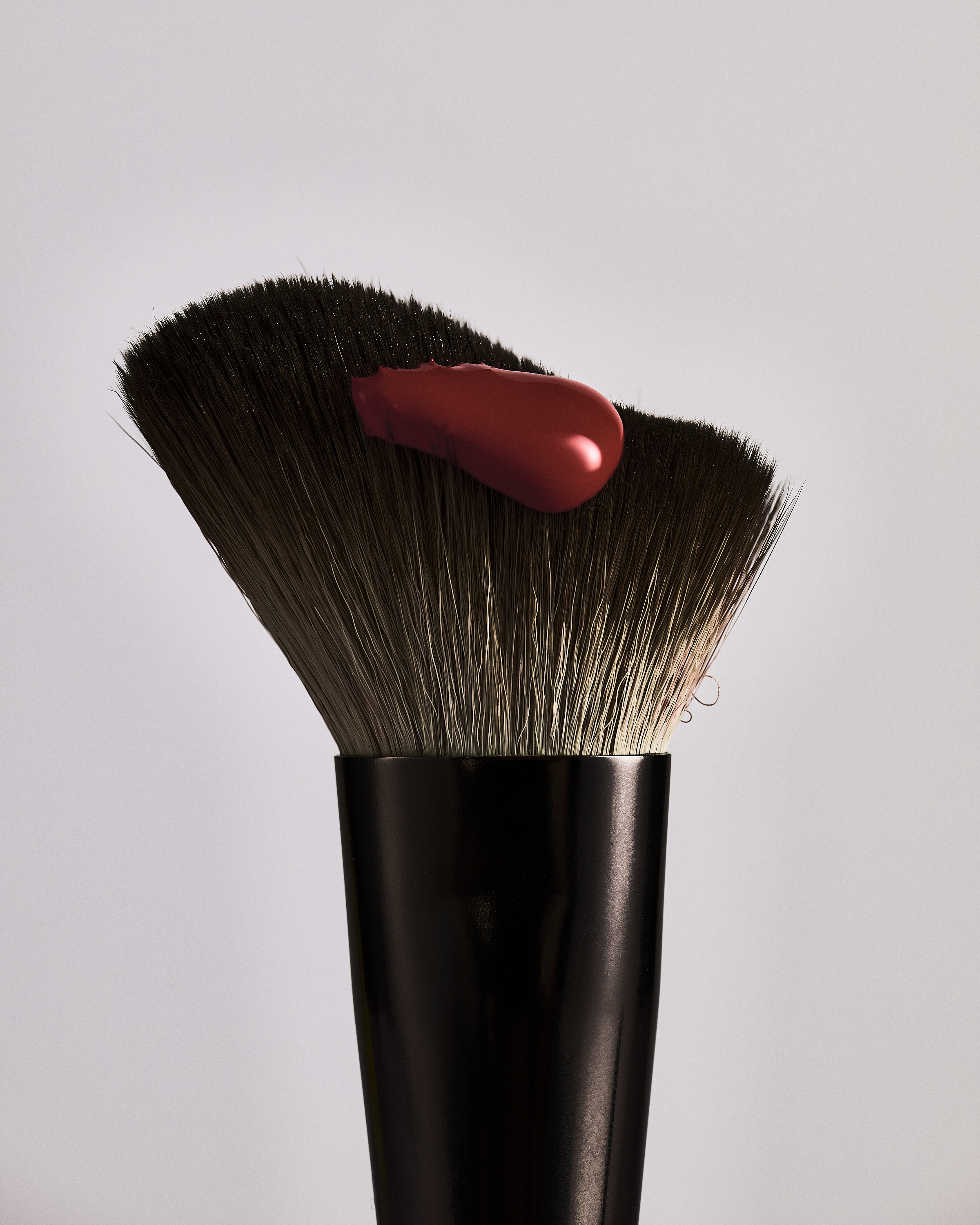 Macro shot of a makeup brush with a vibrant red cosmetic product on its bristles, against a gradient white background, emphasizing beauty textures.