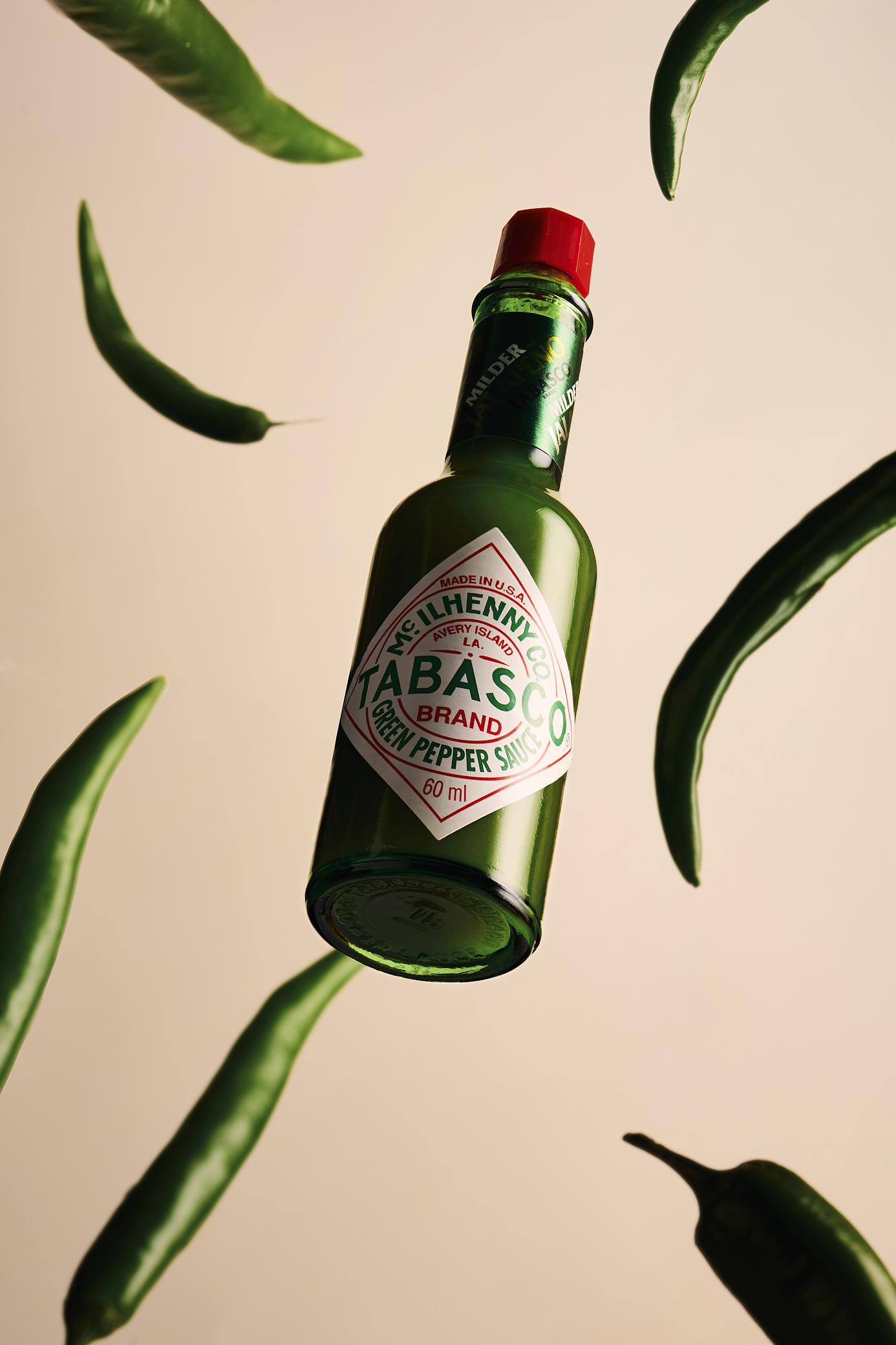 Floating green Tabasco sauce bottle with fresh green chile peppers against a pale background in a product still life.