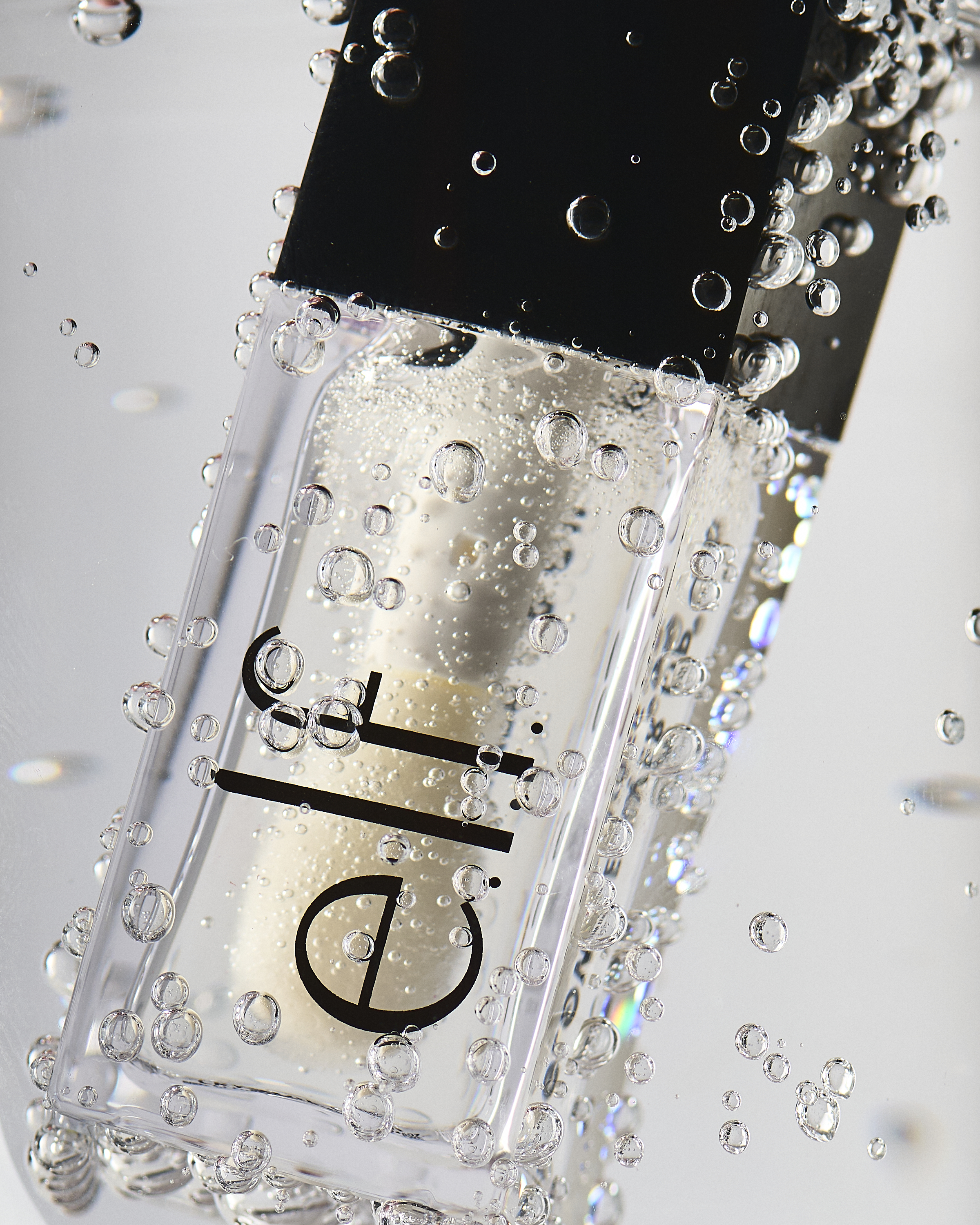 Close-up of a clear ELF lip gloss bottle with a black cap surrounded by water droplets against a bright, clean background.