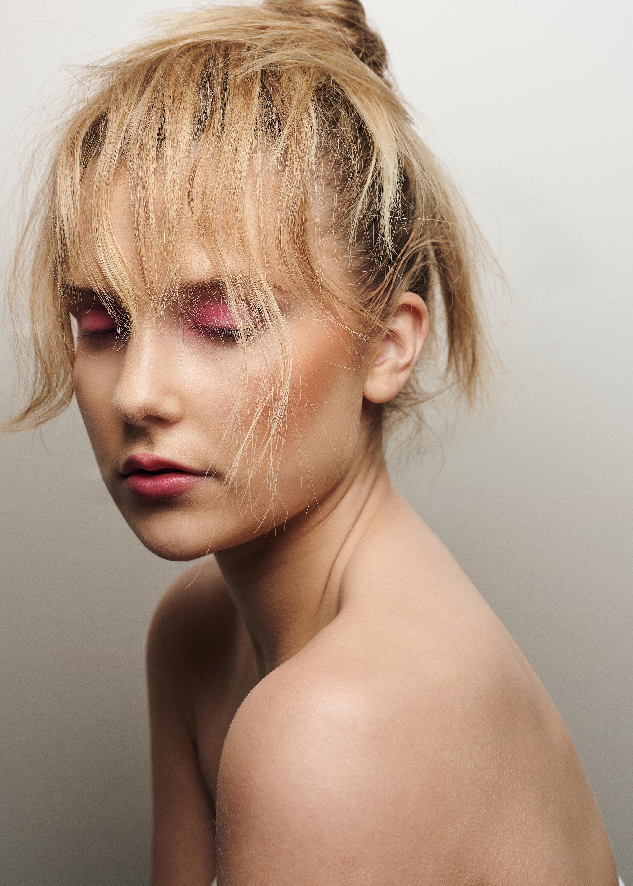 Close-up studio portrait of a blond-haired young adult woman with pink eye shadow and bare shoulders posing against a light grey background.