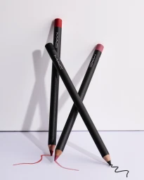 Three MAC lip and eye makeup pencils balanced upright on their tips over drawn cosmetic swatches on a white background.