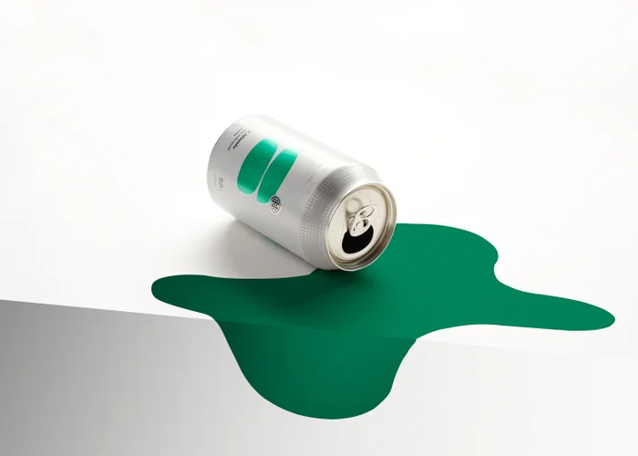 An open silver beverage can resting on its side atop a stylized green paper spill over the edge of a white surface.