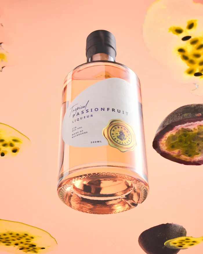 Glass bottle of tropical passionfruit liqueur floating among sliced and halved passionfruit against a soft peach background.