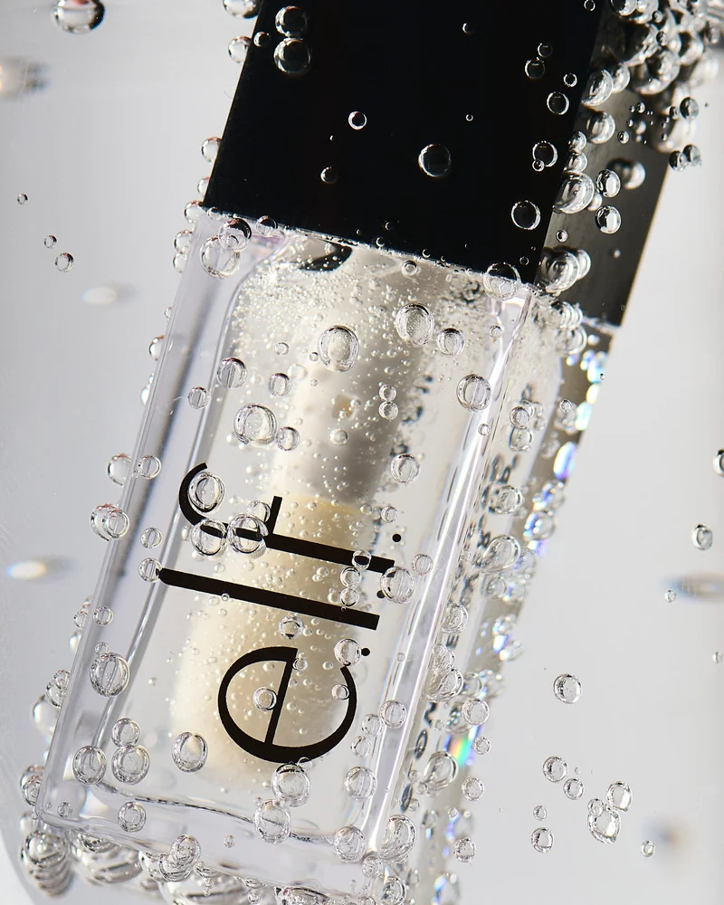Clear e.l.f. lip oil cosmetic bottle with black cap submerged in bubbly water against a bright background.