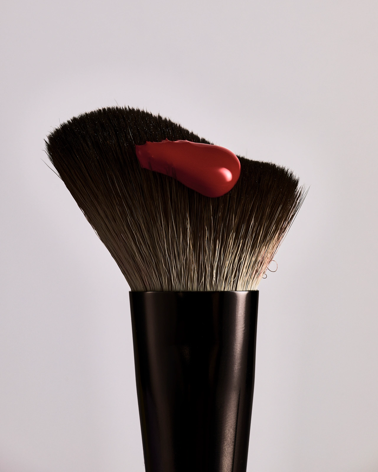 Close-up of a black makeup brush with two-tone bristles topped with a dollop of glossy red cosmetic cream against a plain grey background.