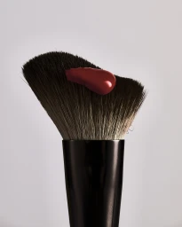 Close-up of a black makeup brush with two-tone bristles topped with a dollop of glossy red cosmetic cream against a plain grey background.
