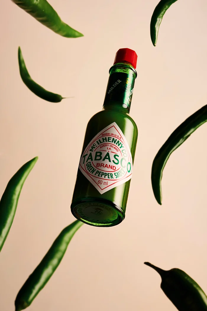 A bottle of Tabasco Green Pepper Sauce suspended mid-air surrounded by floating fresh green chili peppers against a warm neutral backdrop.