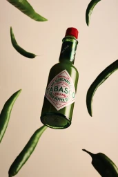 A bottle of Tabasco Green Pepper Sauce suspended mid-air surrounded by floating fresh green chili peppers against a warm neutral backdrop.
