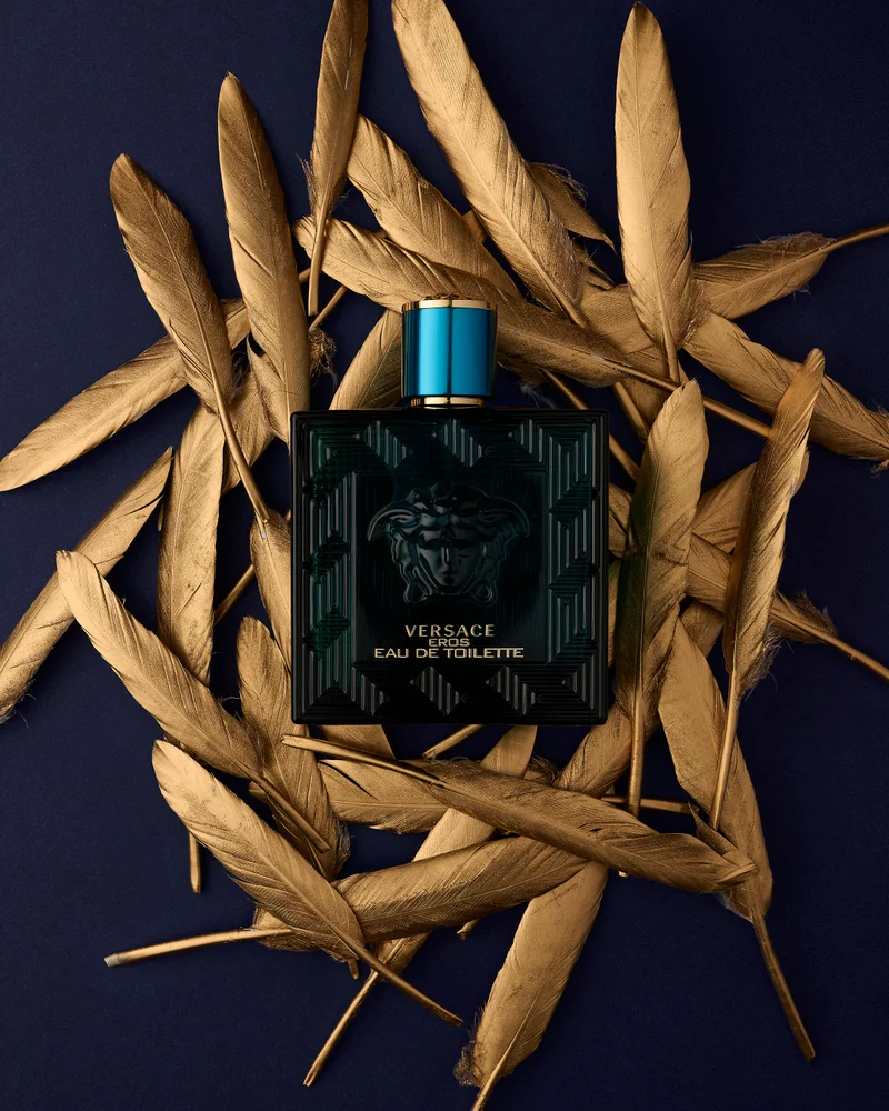 Teal and black fragrance bottle centered within a ring of metallic gold feathers against a dark navy blue background.