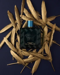 Teal and black fragrance bottle centered within a ring of metallic gold feathers against a dark navy blue background.