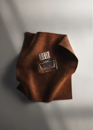 A clear rectangular perfume bottle rests on a draped piece of brown suede over a smooth grey background.