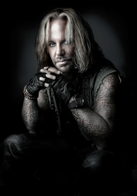 Close-up portrait of a blonde adult male with tattoos wearing fingerless gloves and wrist beads, sitting against a dark background.