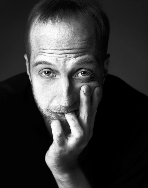 Monochrome portrait of a light-skinned adult male with short hair, looking at the camera with a thoughtful facial expression and hand near face.