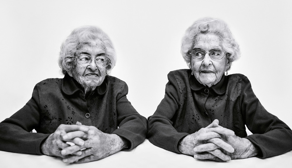 Black and white portrait of two elderly women with short white hair, wearing jackets, and sitting side-by-side with hands clasped.