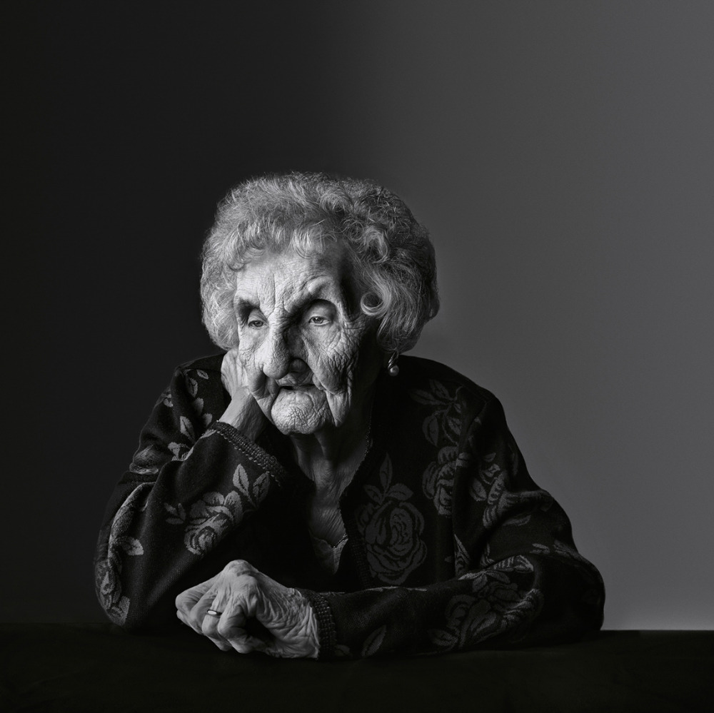 Elderly woman portrait in black and white, resting head on hand at a table, wearing floral patterned top against dark backdrop.