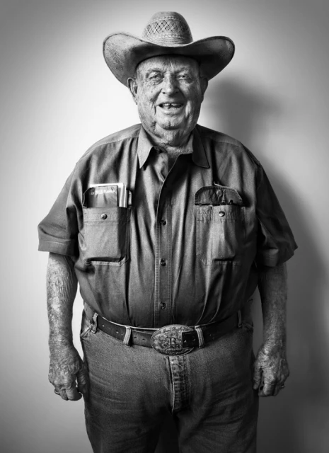 Black and white portrait of an elderly man wearing a cowboy hat, button-down shirt, jeans, and belt with a large buckle, smiling at the camera.