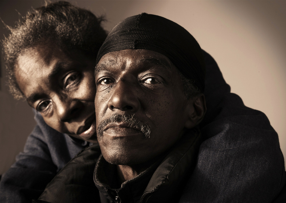 Close-up portrait of an African American couple. The man is wearing a black cap and the woman is behind him, looking at the camera.