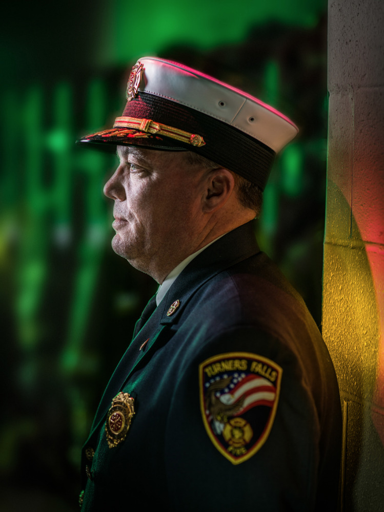 Profile portrait of a firefighter in uniform with a hat, featuring a patch, in an indoor setting with green and yellow lighting.