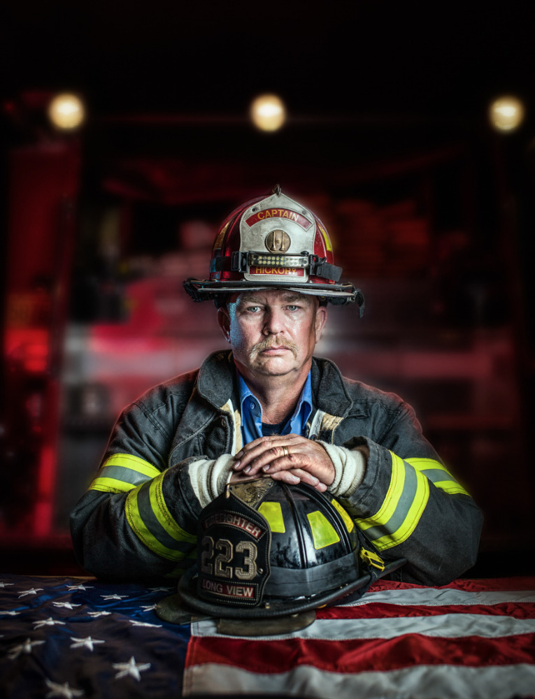 Portrait of a firefighter in uniform with a helmet resting on a US flag, set against a dark background with blurred lights.