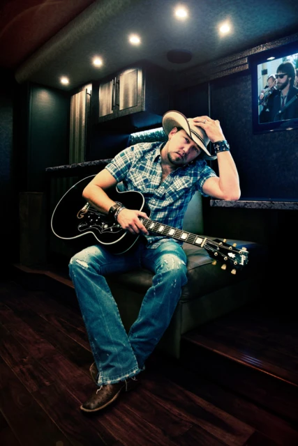 Adult male musician wearing a cowboy hat and holding a guitar while seated inside a vehicle with a TV screen in the background.