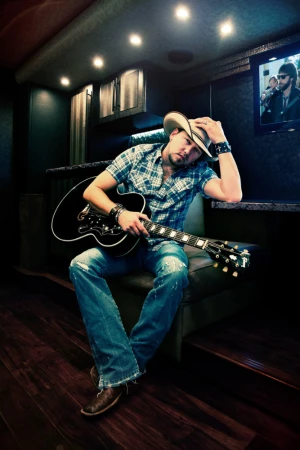 Adult male musician wearing a cowboy hat and holding a guitar while seated inside a vehicle with a TV screen in the background.