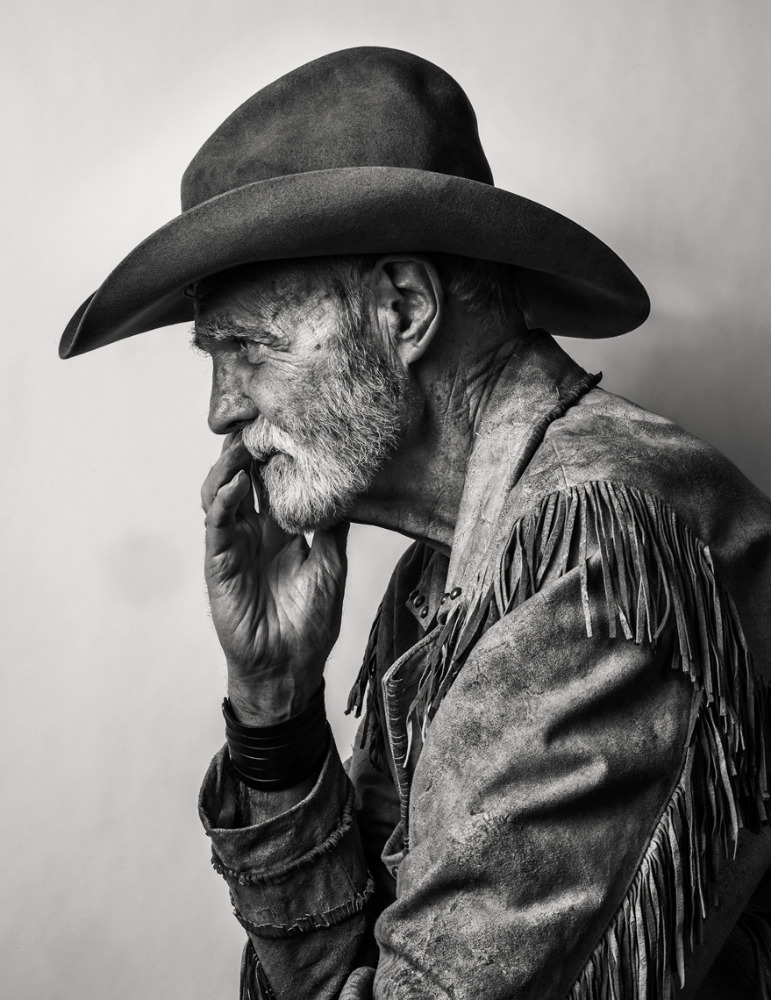 Monochrome portrait of an elderly man with a beard wearing a cowboy hat and fringed jacket, his hand raised to his chin in thought.