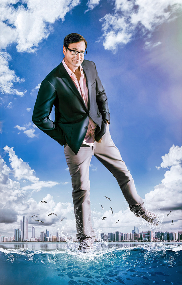 An adult male with glasses and a blazer poses, seeming to stride over a flooded city with a cloudy, blue sky in the background.