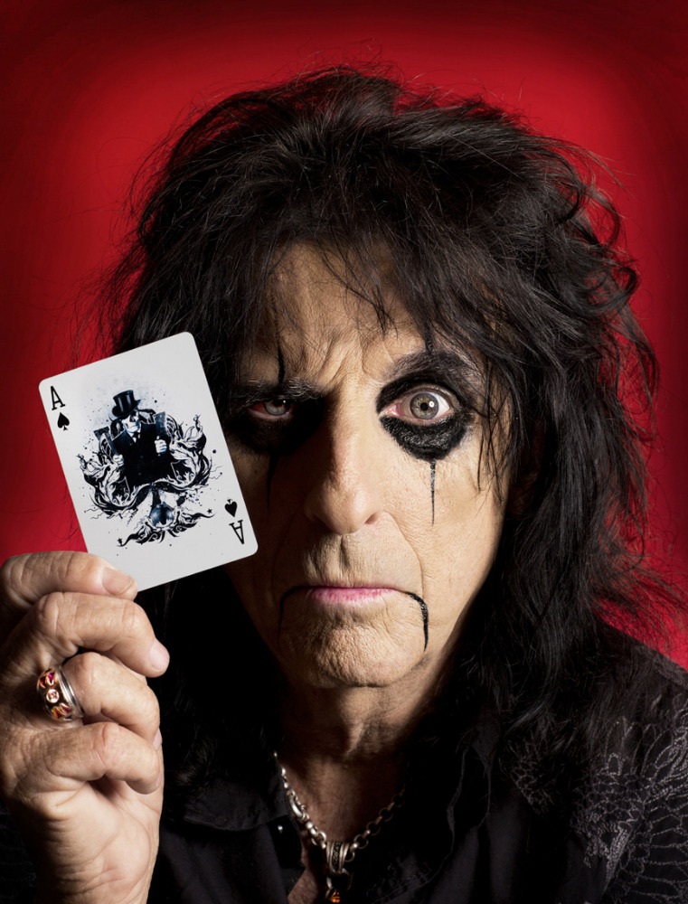 Close-up of Alice Cooper holding an ace playing card with dark makeup and black hair against a red backdrop.