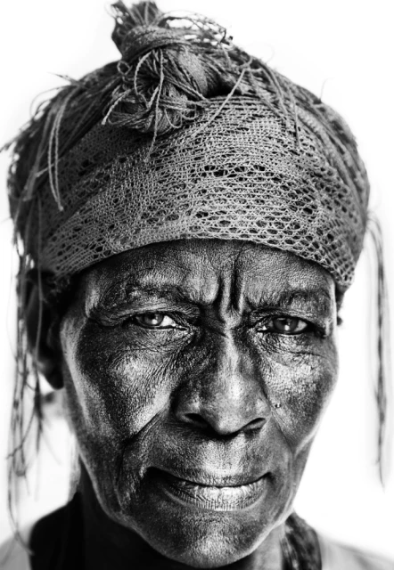 Striking black and white portrait of an elderly Black woman with a wrinkled face and a patterned headscarf, set against a bright white background.