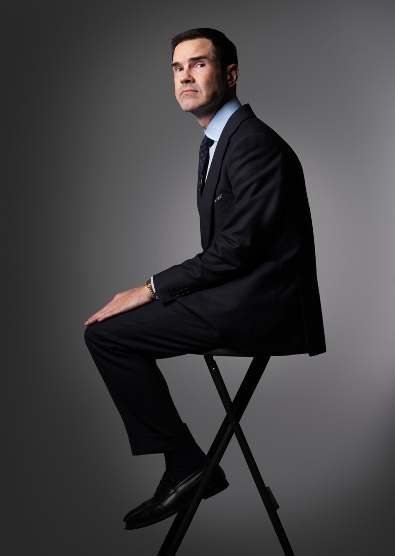 A man in a dark suit sits on a black folding stool against a grey background, looking to the side in a formal studio portrait.