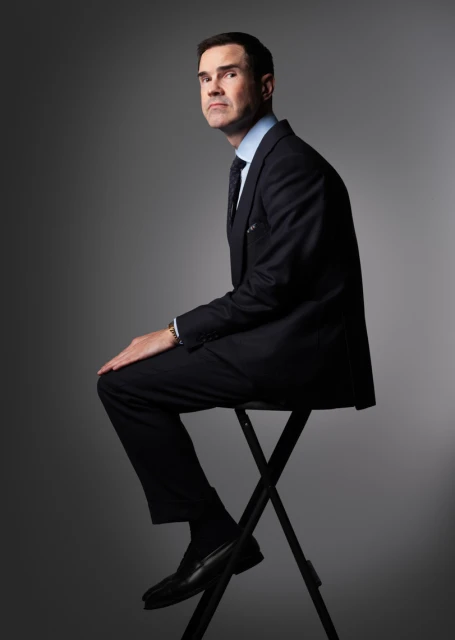A man in a dark suit sits on a black folding stool against a grey background, looking to the side in a formal studio portrait.