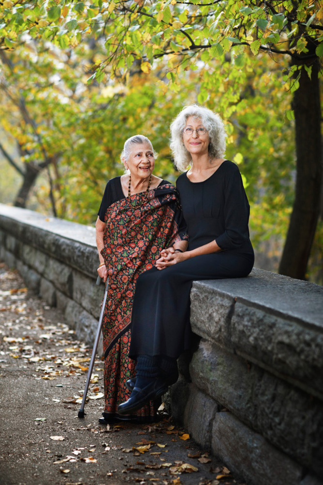 Elderly woman with a cane poses with an adult woman outside in a park on a stone ledge with foliage in the background.
