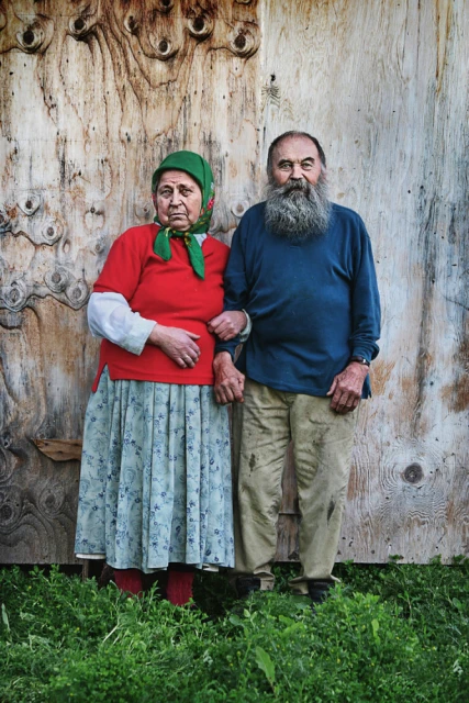 An elderly couple stands outdoors near a textured wall. The woman wears a red top and the man a blue top, both holding hands.