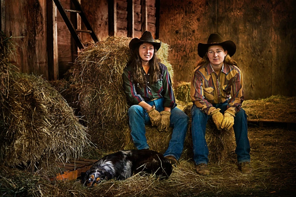 Two women in cowboy hats sit on hay bales with a dog in a rustic barn interior, wearing jeans and gloves, with natural light.