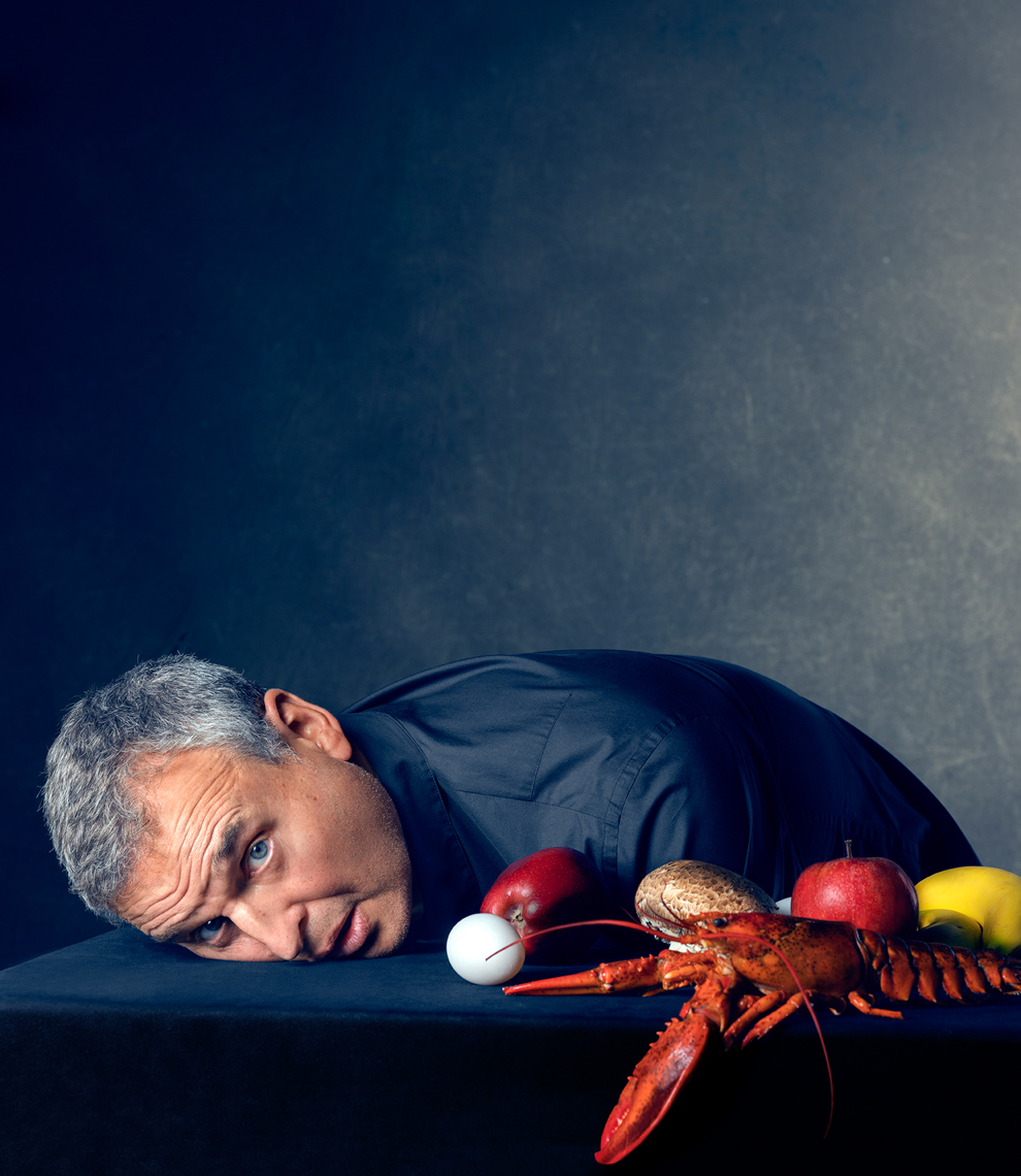Man lays on black surface with apples, lemon, mushroom, golf ball, and lobster in still life composition against dark background.