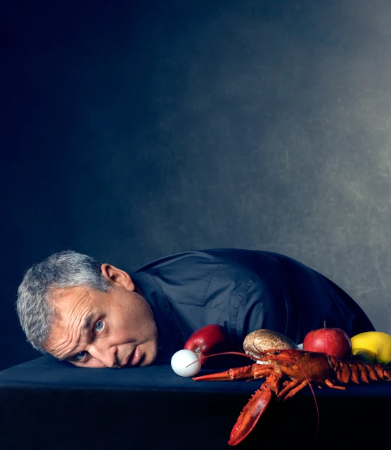Man lays on black surface with apples, lemon, mushroom, golf ball, and lobster in still life composition against dark background.