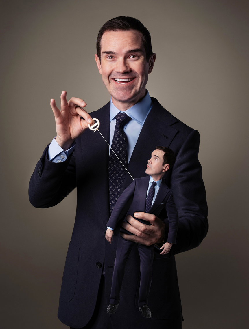 Adult male entertainer holds a puppet version of himself by a string in a studio portrait against a neutral gray background.