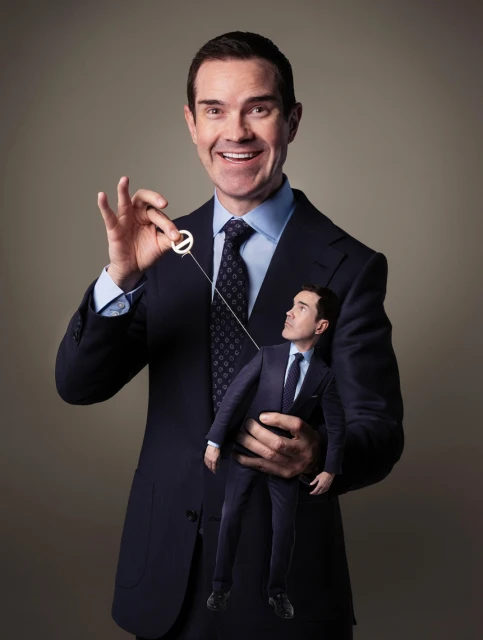 Adult male entertainer holds a puppet version of himself by a string in a studio portrait against a neutral gray background.