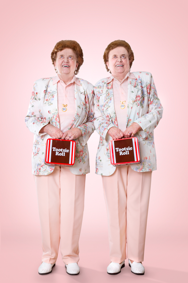Two elderly women in matching floral jackets and pink pants hold Tootsie Roll lunchboxes against a pink background, full-length shot.
