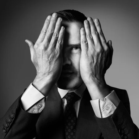 Monochrome portrait of a man in a suit holding his hands up to his face, covering his eyes with his palms.