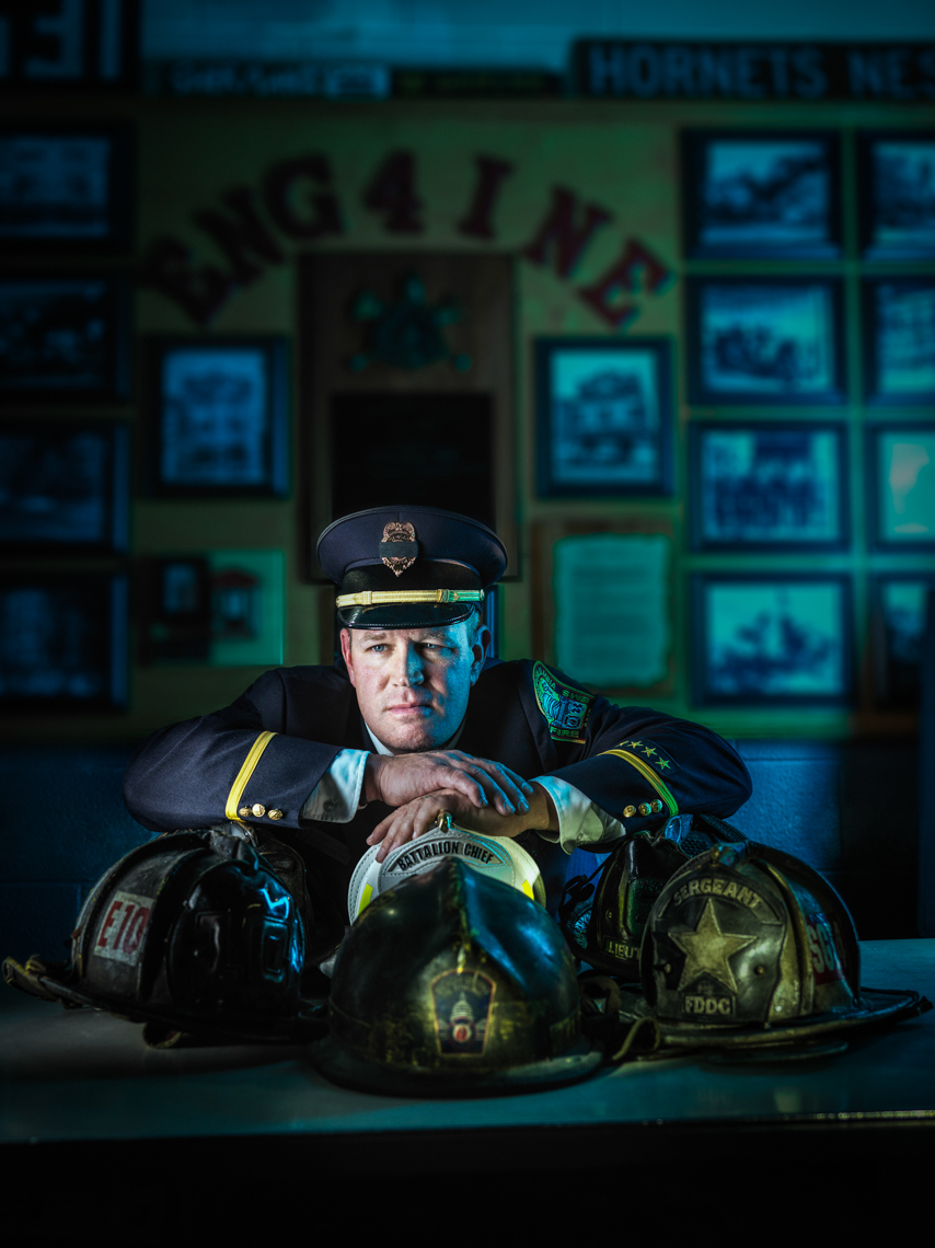 Portrait of a firefighter wearing a uniform and hat, resting his arms on three different fireman helmets against a blurred backdrop.