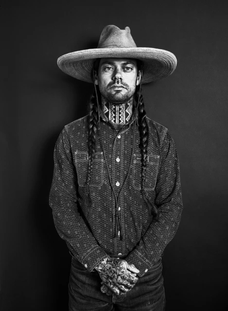 Monochrome portrait of a man in a sombrero, collared shirt, and jeans, standing against a dark background, showcasing his tattoos and braids.